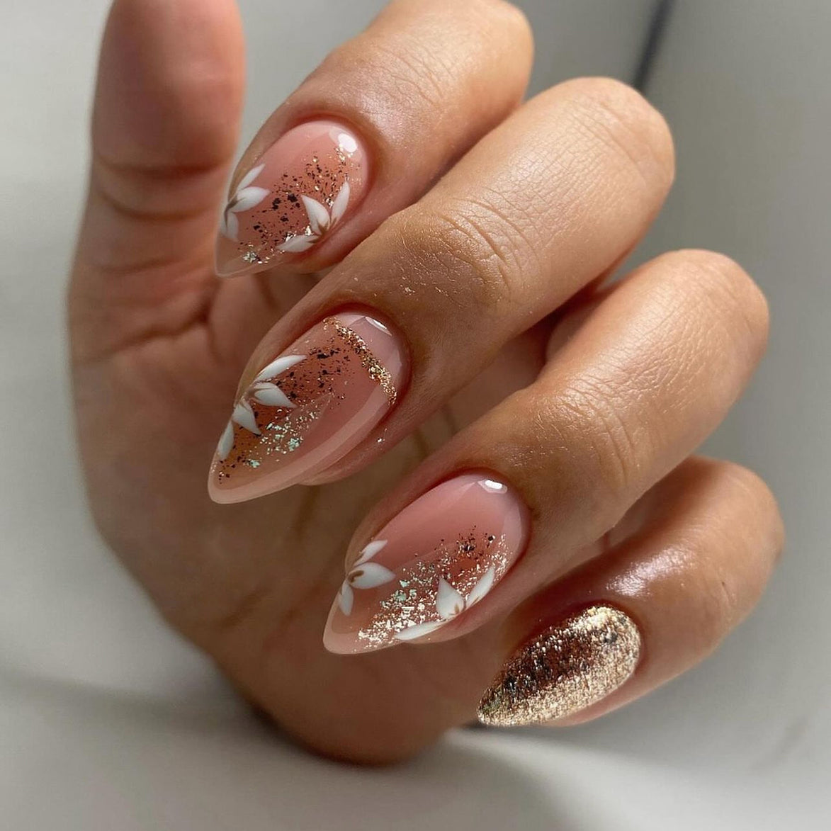 Hand wearing peach almond press-on nails with white floral art and rose gold glitter tips