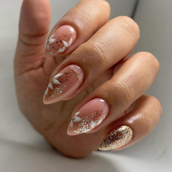 Hand wearing peach almond press-on nails with white floral art and rose gold glitter tips